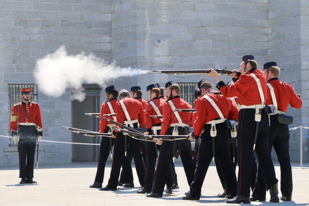 Historical reenactors in red and black uniforms firing muskets in formation at Fort Henry courtyard.